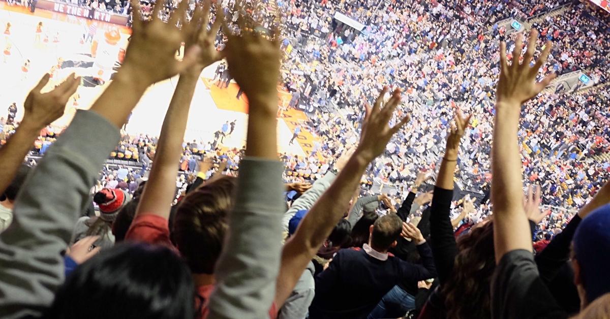 An overhead view of a packed indoor basketball arena with raised arms of excited spectators in the foreground and a brightly lit court in the background.