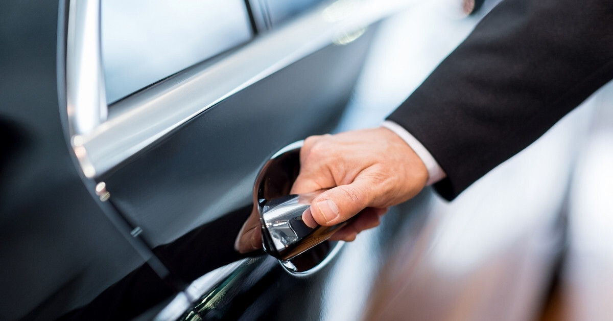 A close-up of a hand in formal attire opening the chrome door handle of a black luxury car, with a blurred background suggesting a professional or service setting.