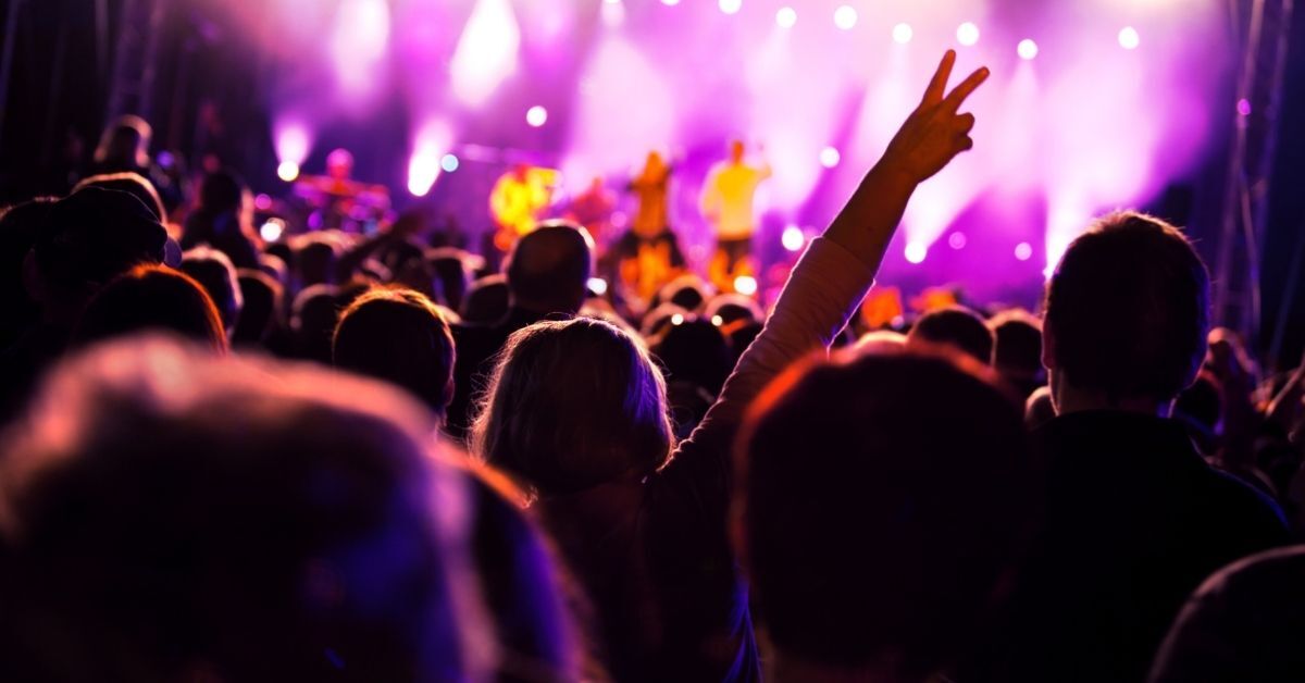 A vibrant scene of a live concert with a crowd of people raising their hands in excitement, silhouetted against a brightly lit stage with purple and pink lighting.
