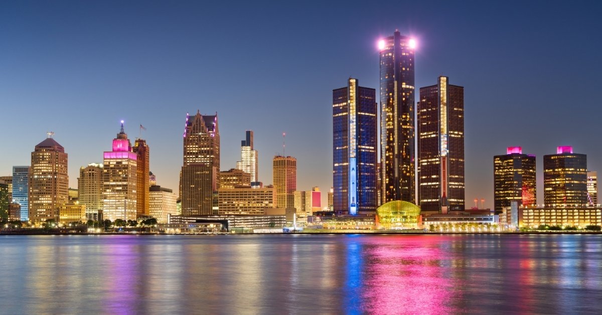 A panoramic nighttime view of the Detroit skyline across a river, featuring the illuminated Renaissance Center and colorful reflections on the water.