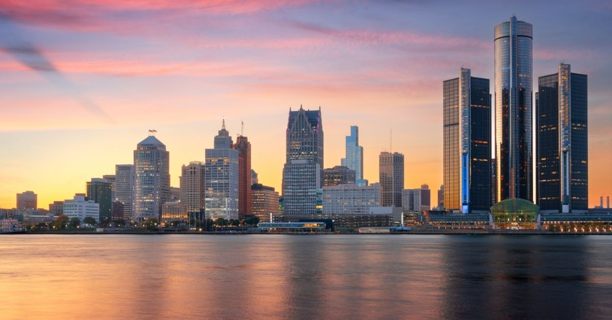A panoramic view of the Detroit city skyline at sunset, featuring the Renaissance Center and other skyscrapers, with colorful reflections on a calm body of water.