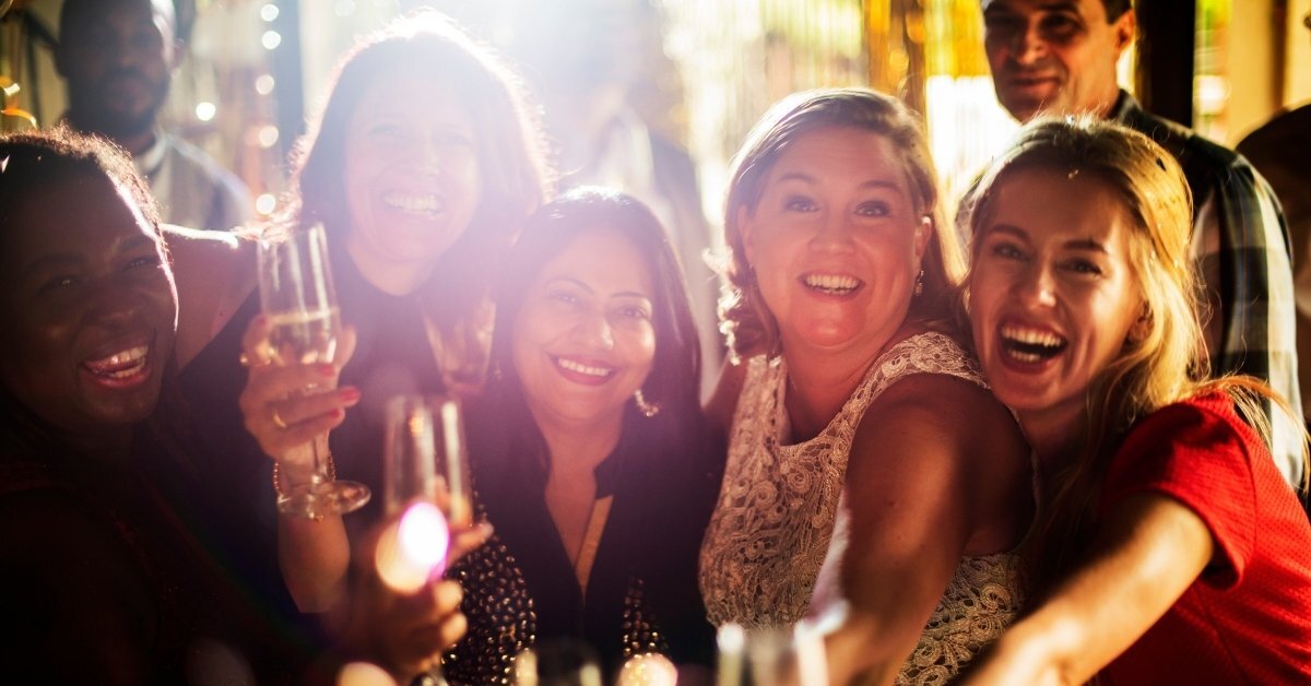 A group of six friends, predominantly women, enjoying a festive indoor gathering with champagne flutes, warm lighting, and a celebratory atmosphere.